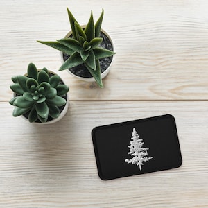 May include: A black fabric patch with a white embroidered pine tree design. The patch is on a white wooden surface with two potted succulents.