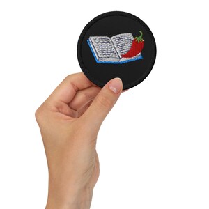 May include: A round, black embroidered patch featuring an open book with blue trim and a red chili pepper. The patch is held in a hand, showcasing the detailed stitching and vibrant colors of the design.