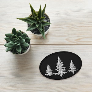 May include: Black oval patch featuring three white embroidered pine trees. The patch is set against a light wood grain surface, with two small potted succulents in the background.