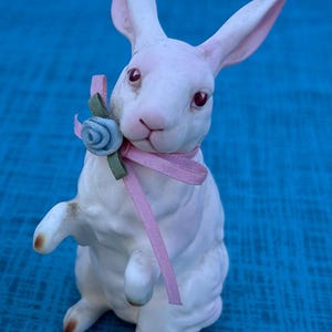 May include: A white ceramic rabbit figurine with pink-lined ears and red eyes. It has a pink ribbon and a blue flower around its neck. The rabbit is posed standing upright, with one paw raised.