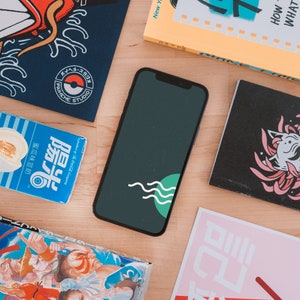 May include: A smartphone with a dark green screen featuring a white wave design and a green circle, surrounded by books and packaging. The items are on a light wood surface. Some items have Japanese text.