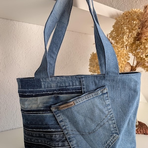 Könnte beinhalten: Eine blaue Jeans-Tragetasche mit Patchwork-Design. Die Tasche hat eine Vordertasche aus einer recycelten Jeans-Tasche. Die Tasche hat zwei Griffe und besteht aus verschiedenen Blautönen aus Jeansstoff.