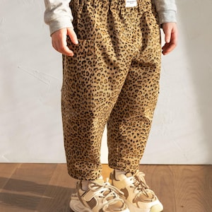 May include: Tan and brown leopard print pants with pockets. The pants are worn with a gray long-sleeve shirt and beige and white sneakers. The pants have a tag that says "DEAR KELLY".