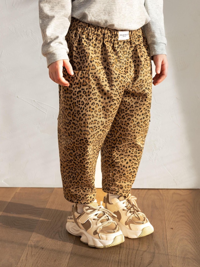 May include: Tan and brown leopard print pants with pockets. The pants are worn with a gray long-sleeve shirt and beige and white sneakers. The pants have a tag that says "DEAR KELLY".