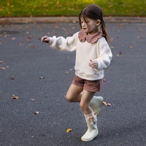 May include: A young child wearing a white sweater with a red and white checkered collar, matching shorts, and white boots with bows. The child is outside on a paved surface with fallen leaves.