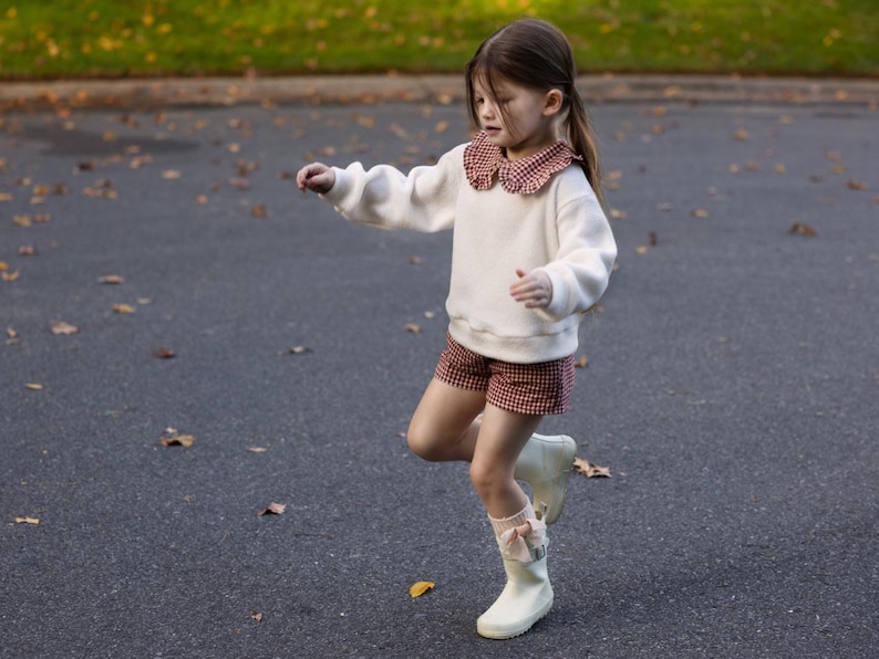 May include: A young child wearing a white sweater with a red and white checkered collar, matching shorts, and white boots with bows. The child is outside on a paved surface with fallen leaves.