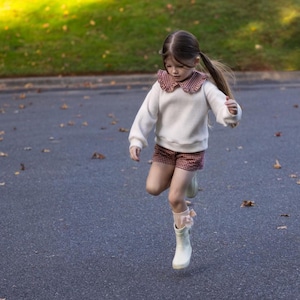 May include: A young child wearing a white sweater with a red and white patterned collar and matching shorts. The child is wearing white boots and socks with pink bows, jumping on a paved surface.