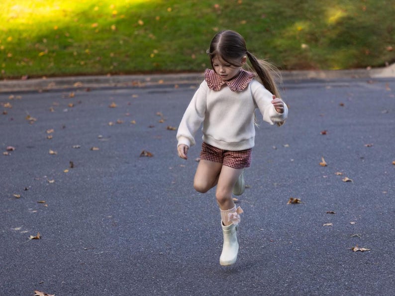 May include: A young child wearing a white sweater with a red and white patterned collar and matching shorts. The child is wearing white boots and socks with pink bows, jumping on a paved surface.