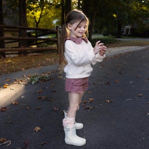 May include: A young person wearing a white sweater with a red and white checkered collar and matching shorts. They are wearing white boots with pink bows. The outfit is suitable for a child's fashion or a fall season look.