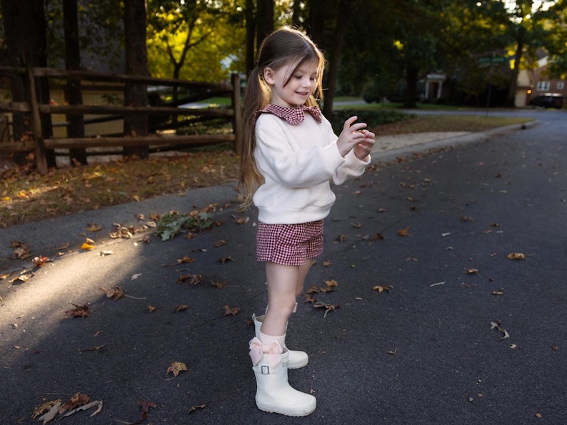 May include: A young person wearing a white sweater with a red and white checkered collar and matching shorts. They are wearing white boots with pink bows. The outfit is suitable for a child's fashion or a fall season look.