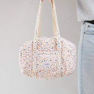 May include: A quilted floral print tote bag with cream-colored handles and a front pocket. The bag features a pattern of small blue, red, and yellow flowers on a white background. The text "DEAR BILLY" is visible on the front pocket.