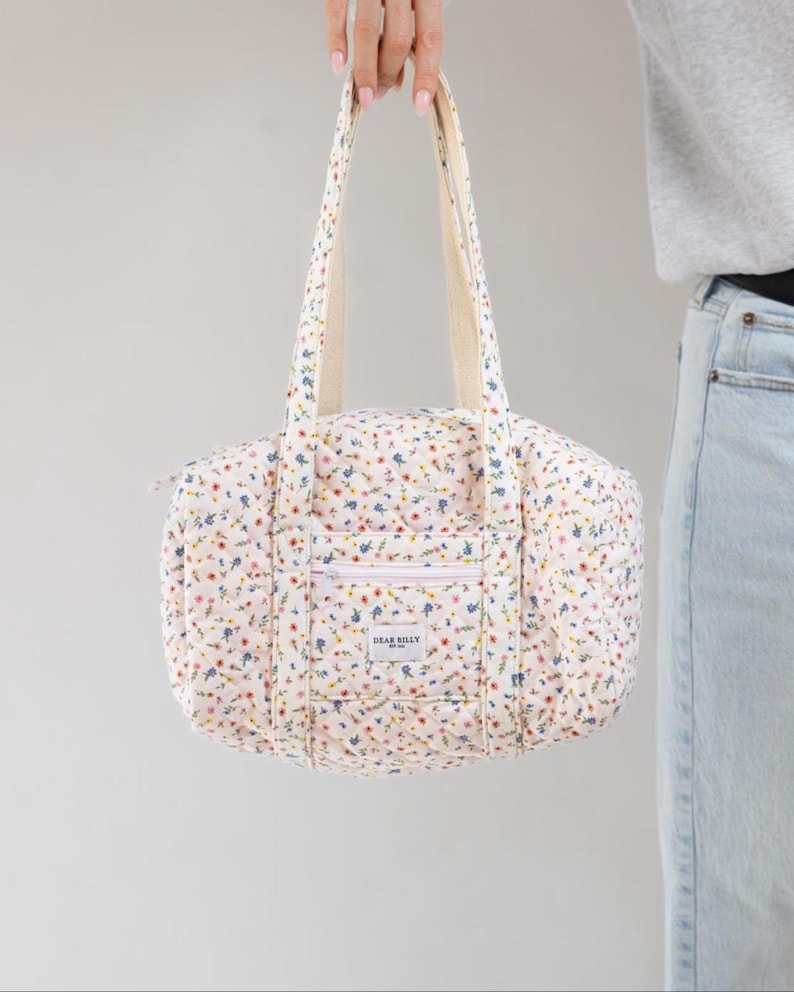 May include: A quilted floral print tote bag with cream-colored handles and a front pocket. The bag features a pattern of small blue, red, and yellow flowers on a white background. The text "DEAR BILLY" is visible on the front pocket.
