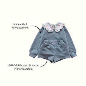 May include: A light blue Honor Roll sweatshirt and matching GRWM boxer shorts. The sweatshirt has a floral-patterned collar. The shorts have pockets and a small label. The set is made of a soft material.