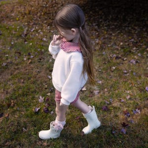 May include: A young child wearing a white sweater with a red and white checkered collar and matching shorts. The child is also wearing white rain boots and pink socks with bows. The child is walking on a grassy area with fallen leaves.
