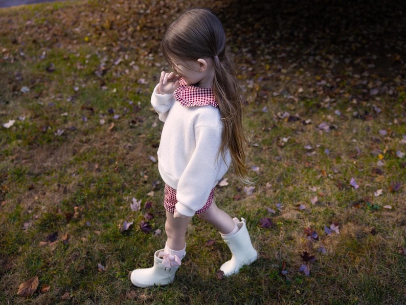 May include: A young child wearing a white sweater with a red and white checkered collar and matching shorts. The child is also wearing white rain boots and pink socks with bows. The child is walking on a grassy area with fallen leaves.