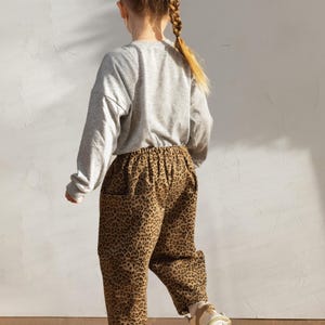 May include: A child wearing a gray long-sleeved shirt and brown leopard print pants. The child is also wearing white and tan sneakers. The outfit is casual and suitable for everyday wear.