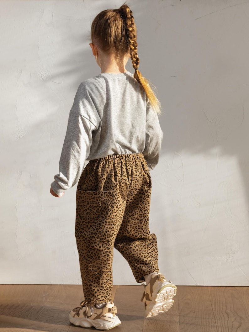 May include: A child wearing a gray long-sleeved shirt and brown leopard print pants. The child is also wearing white and tan sneakers. The outfit is casual and suitable for everyday wear.