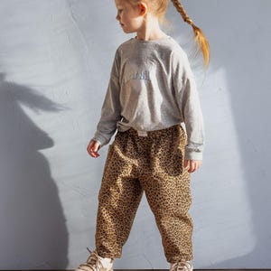 May include: A young person wearing a gray long-sleeve shirt with the text "dear dill" and leopard print pants. The person is also wearing beige sneakers. The outfit is casual and suitable for everyday wear.