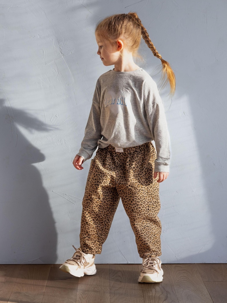 May include: A young person wearing a gray long-sleeve shirt with the text "dear dill" and leopard print pants. The person is also wearing beige sneakers. The outfit is casual and suitable for everyday wear.