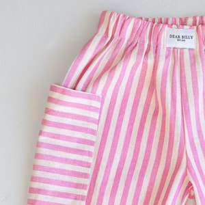 May include: Close-up of pink and white striped pants with a pocket. The pants have an elastic waistband and a tag that reads "DEAR BILLY EST. 2021". The fabric has a linen-like texture.