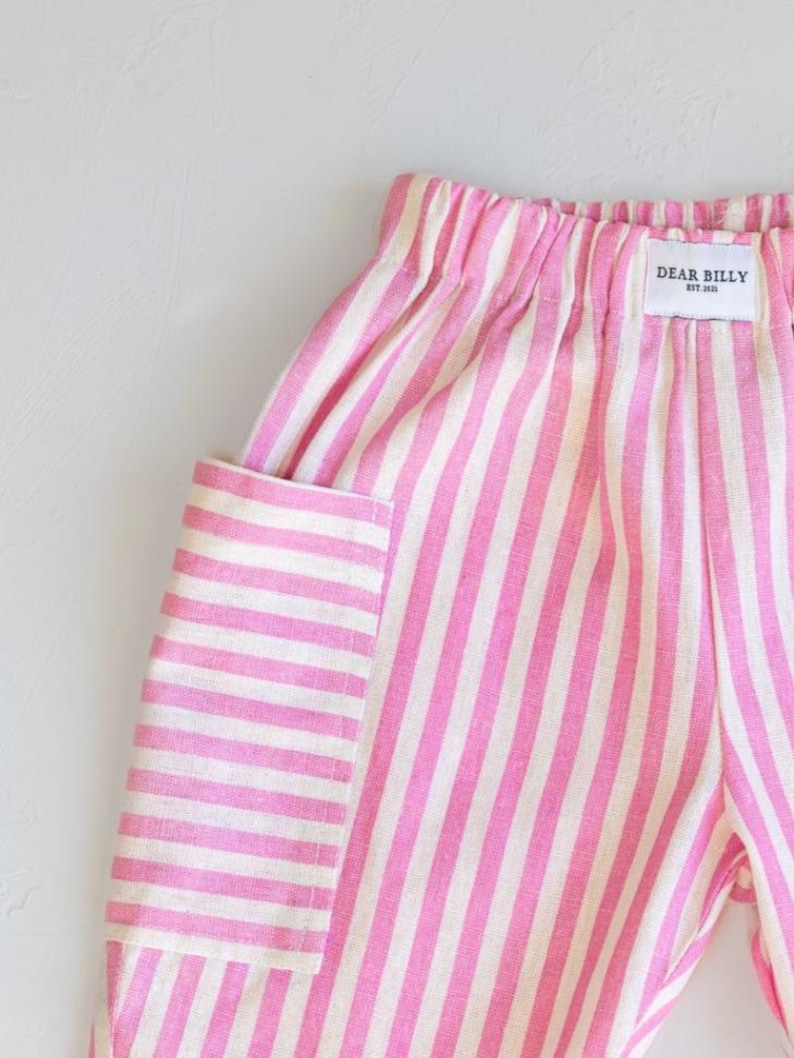 May include: Close-up of pink and white striped pants with a pocket. The pants have an elastic waistband and a tag that reads "DEAR BILLY EST. 2021". The fabric has a linen-like texture.