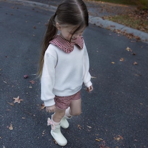 May include: A young person wearing a white sweatshirt with a red and white checkered collar and matching shorts. They are also wearing white boots with pink bows. The outfit is suitable for a child.