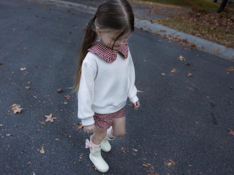 May include: A young person wearing a white sweatshirt with a red and white checkered collar and matching shorts. They are also wearing white boots with pink bows. The outfit is suitable for a child.
