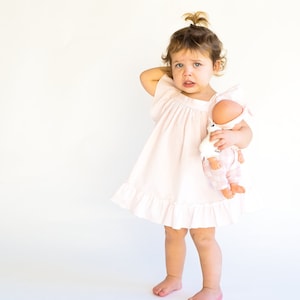 May include: A young child in a pink dress holds a doll with a pink and white outfit. The child is looking at the doll with a serious expression.