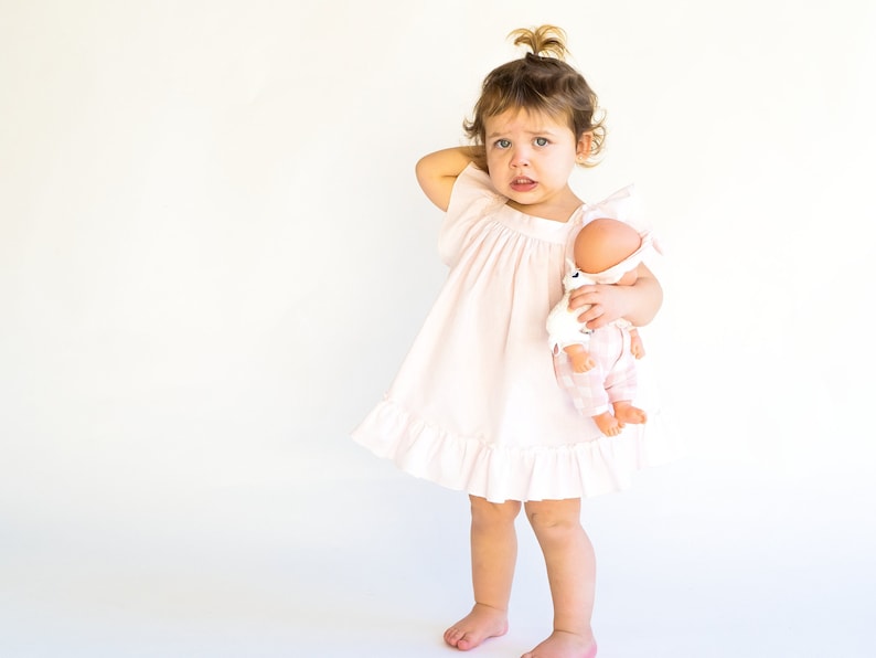 May include: A young child in a pink dress holds a doll with a pink and white outfit. The child is looking at the doll with a serious expression.