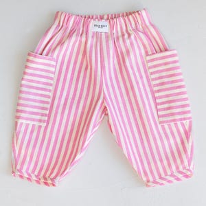 May include: Pink and white striped baby pants with two large pockets. The pants have an elastic waistband and the text "DEAR BILLY" is printed on the back of the waistband. The pants are made of a soft fabric.