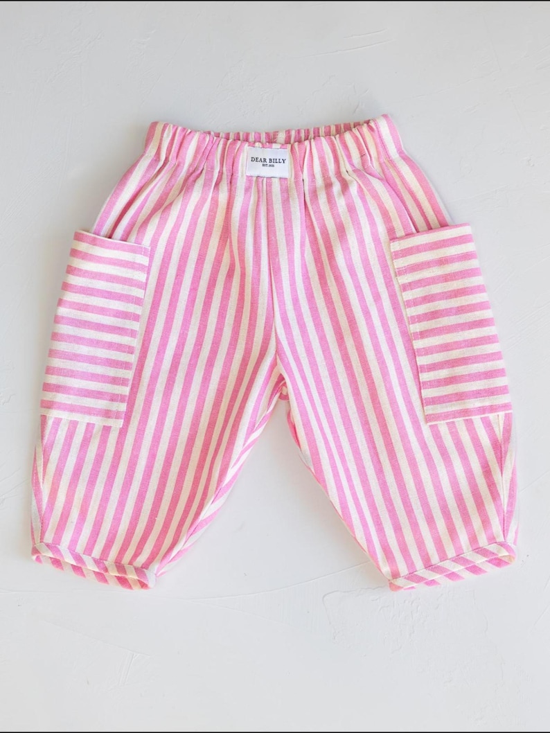 May include: Pink and white striped baby pants with two large pockets. The pants have an elastic waistband and the text "DEAR BILLY" is printed on the back of the waistband. The pants are made of a soft fabric.