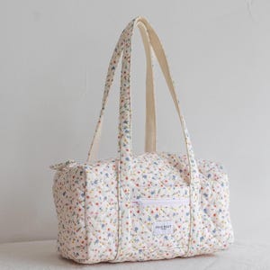 May include: A quilted floral print duffel bag with cream-colored handles and a white zipper pocket. The bag features a pattern of small blue, pink, and yellow flowers on a white background. The text "DEAR BILLY" is visible on the pocket.