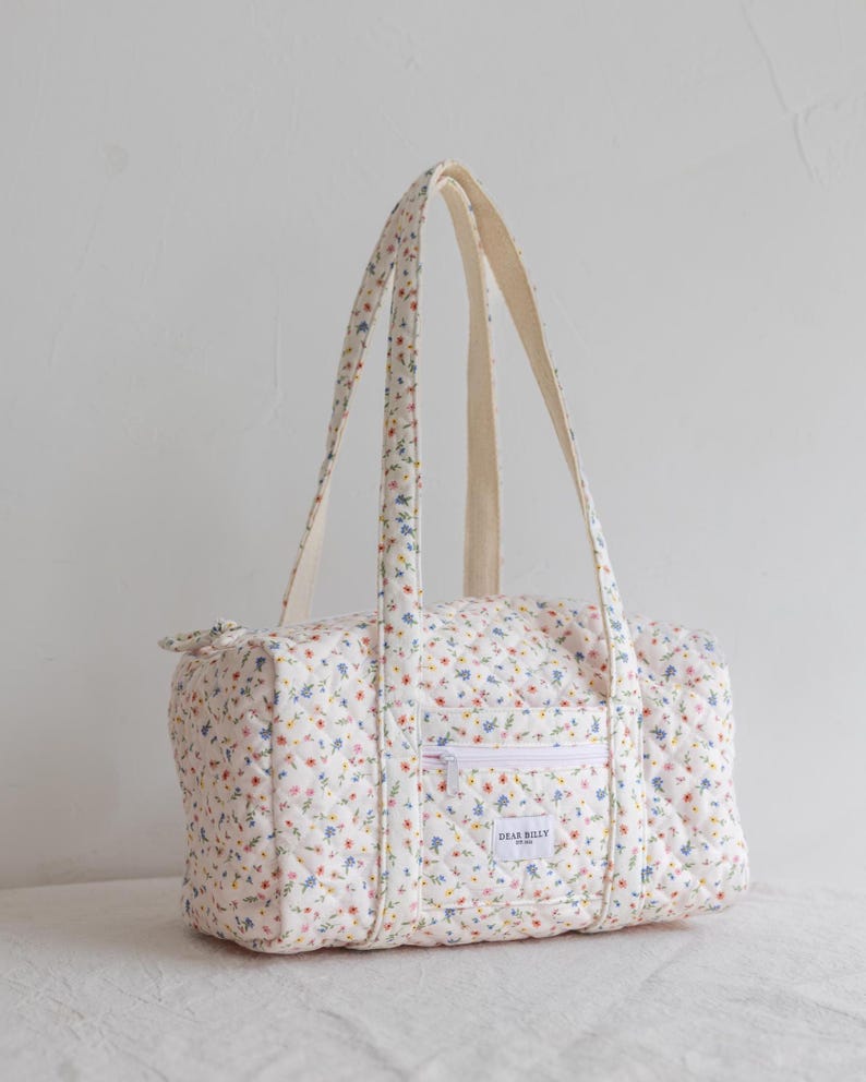 May include: A quilted floral print duffel bag with cream-colored handles and a white zipper pocket. The bag features a pattern of small blue, pink, and yellow flowers on a white background. The text "DEAR BILLY" is visible on the pocket.