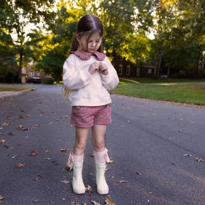 May include: A young person stands on a paved road, wearing a white sweater with a red and white checkered collar, matching shorts, and white boots with pink bows. The background features trees and houses.
