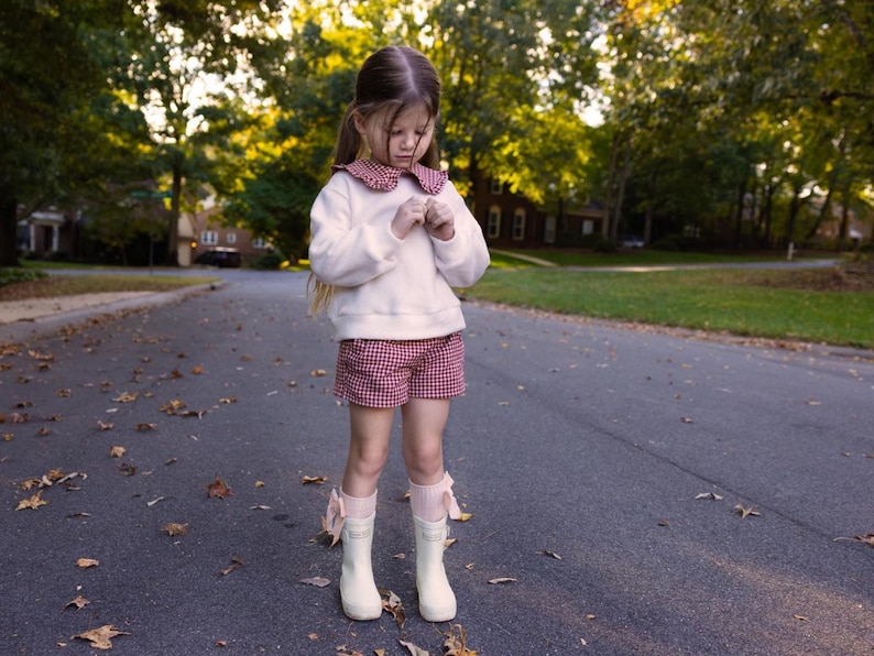 May include: A young person stands on a paved road, wearing a white sweater with a red and white checkered collar, matching shorts, and white boots with pink bows. The background features trees and houses.