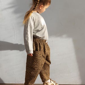 May include: A child wearing a gray sweatshirt and leopard print pants. The pants are brown with black spots. The child is also wearing beige and white sneakers. The child has a long braid.
