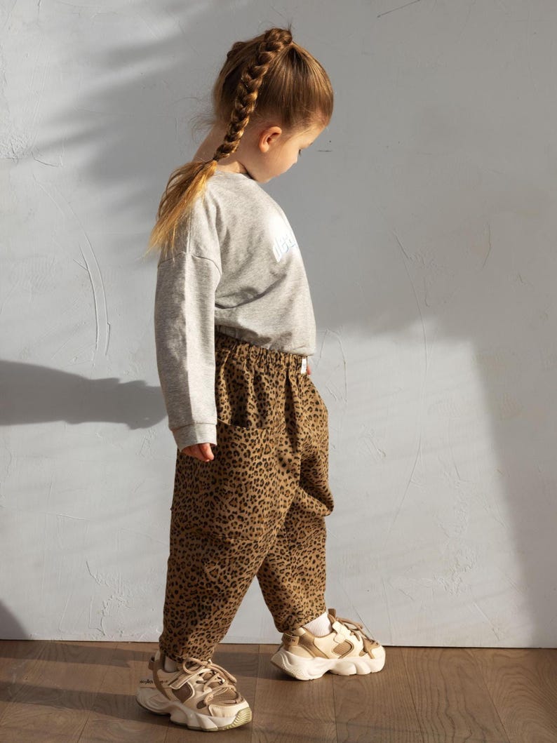 May include: A child wearing a gray sweatshirt and leopard print pants. The pants are brown with black spots. The child is also wearing beige and white sneakers. The child has a long braid.