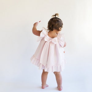 May include: A little girl wearing a pink ruffled dress with a bow in the back.