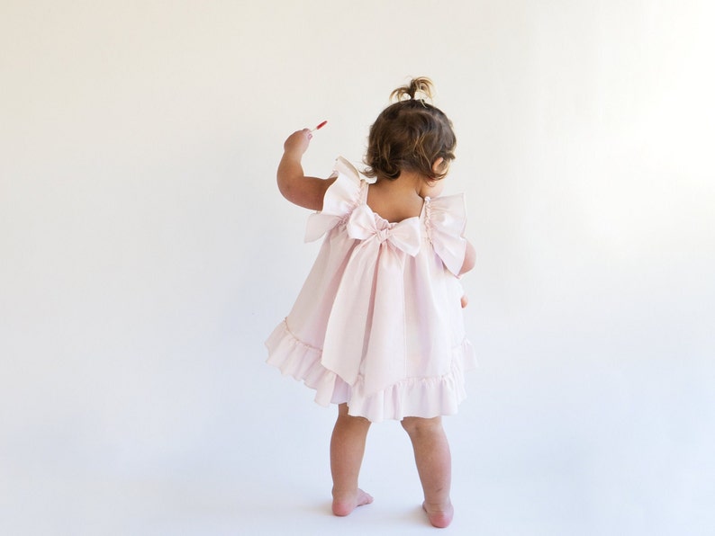 May include: A little girl wearing a pink ruffled dress with a bow in the back.
