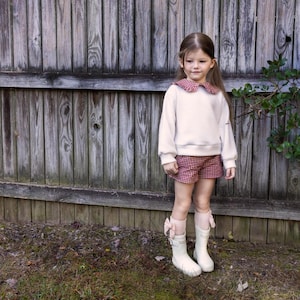 May include: A child's outfit featuring a cream-colored sweater with a red and white gingham collar, paired with matching shorts. The ensemble is completed with white boots and pink bow accents. The outfit is set against a wooden fence background.
