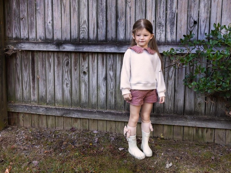 May include: A child's outfit featuring a cream-colored sweater with a red and white gingham collar, paired with matching shorts. The ensemble is completed with white boots and pink bow accents. The outfit is set against a wooden fence background.
