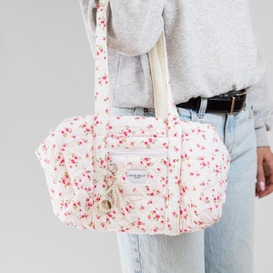 May include: A white quilted tote bag with a floral pattern of small pink roses. The bag has a zipper pocket, two handles, and a small charm. The text "DEAR BILLY" is visible on the front of the bag.