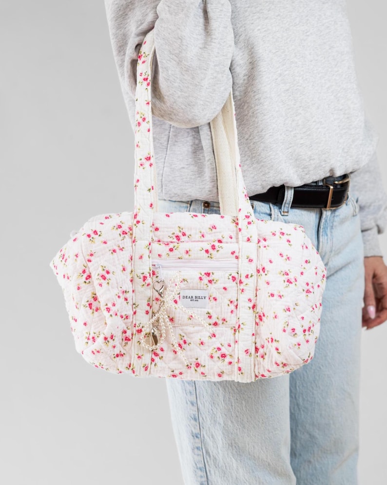 May include: A white quilted tote bag with a floral pattern of small pink roses. The bag has a zipper pocket, two handles, and a small charm. The text "DEAR BILLY" is visible on the front of the bag.
