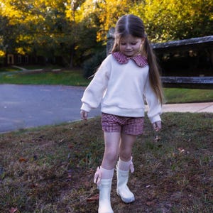 May include: A young person wearing a white sweatshirt with a red and white checkered collar, matching shorts, and white rain boots with pink bows. The outfit is set against a backdrop of fall foliage and a wooden fence.