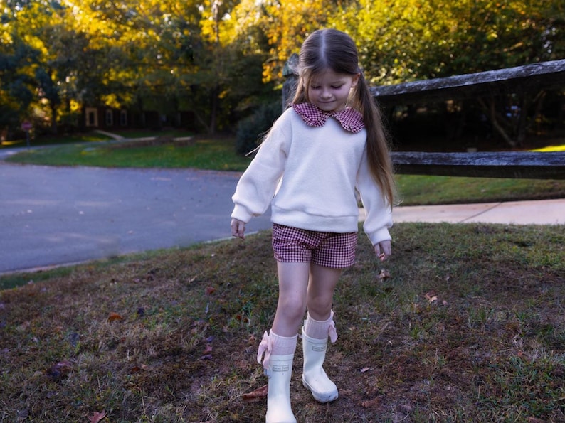May include: A young person wearing a white sweatshirt with a red and white checkered collar, matching shorts, and white rain boots with pink bows. The outfit is set against a backdrop of fall foliage and a wooden fence.