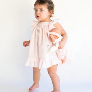 May include: A young child in a pink ruffled dress holds a doll with a pink and white checkered dress. The child is standing on a white background.
