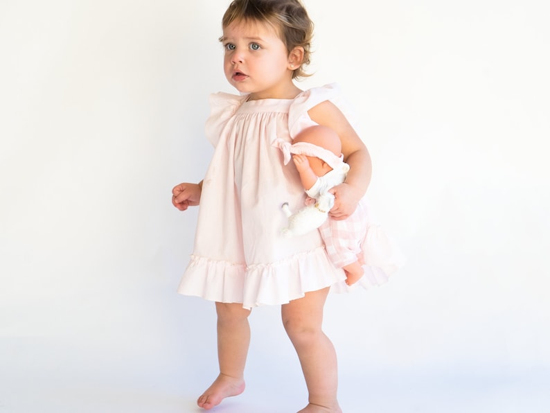 May include: A young child in a pink ruffled dress holds a doll with a pink and white checkered dress. The child is standing on a white background.