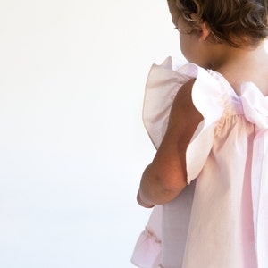 May include: A little girl wearing a pink dress with a large bow in the back. The dress has ruffles on the sleeves and a gathered skirt.
