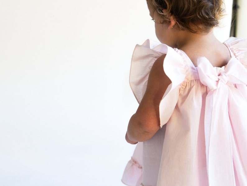 May include: A little girl wearing a pink dress with a large bow in the back. The dress has ruffles on the sleeves and a gathered skirt.