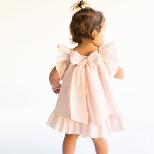 May include: A little girl wearing a pink dress with a large bow on the back. The dress has ruffled sleeves and a ruffled hem.